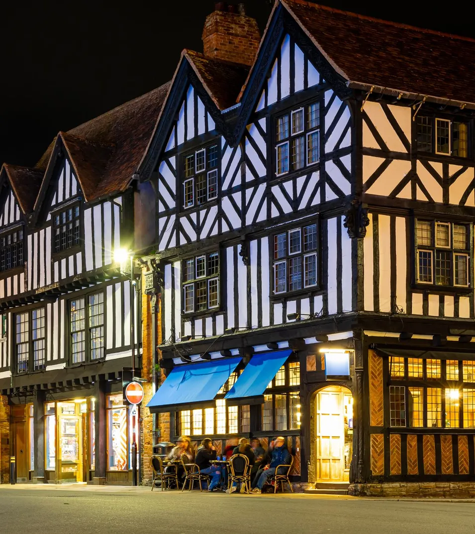 Charming Tudor-style buildings illuminated at night in a festive town square, adorned with hanging lights and holiday decorations, with people dining outside.