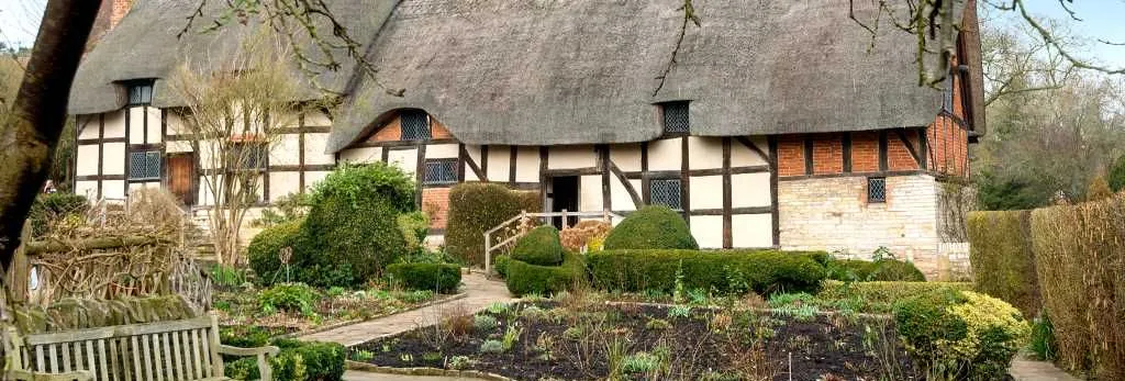Charming thatched-roof cottage with timber framing and brick details, surrounded by a garden with pathways, benches, and trimmed hedges on a clear day.