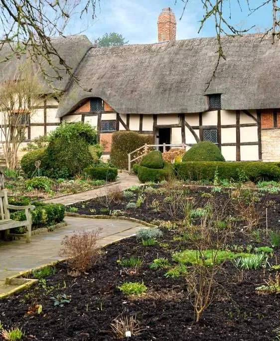 Charming thatched-roof cottage with timber framing and brick details, surrounded by a garden with pathways, benches, and trimmed hedges on a clear day.