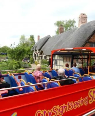 Open-top red City Sightseeing bus in Stratford-upon-Avon, with passengers viewing a charming thatched cottage and lush greenery on a scenic tour.