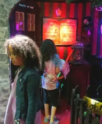 Children exploring a dimly lit, whimsical room filled with Halloween-themed decorations, including a pumpkin with 'Boo!' sign, costumes, and eerie props.