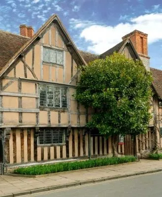 Historic Tudor-style building with timber framing and brick chimneys, surrounded by greenery, under a bright blue sky with scattered clouds.