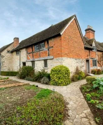 Tudor-style farmhouse with exposed timber and brickwork, surrounded by well-maintained gardens and stone pathways under a clear blue sky.