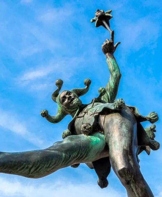 Bronze statue of a playful jester in an animated pose, holding a scepter with a miniature jester head, set against a bright blue sky.