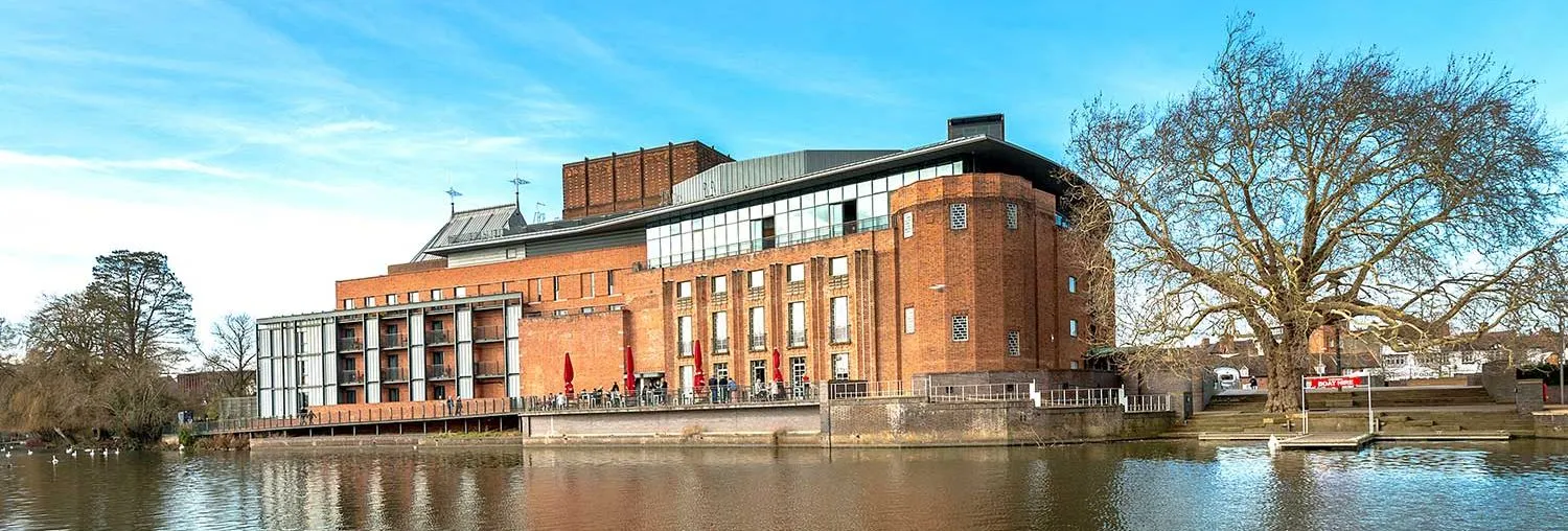 Exterior view of the Royal Shakespeare Theatre, a modern red-brick building located along the riverside, with a bright blue sky and reflections on the water.