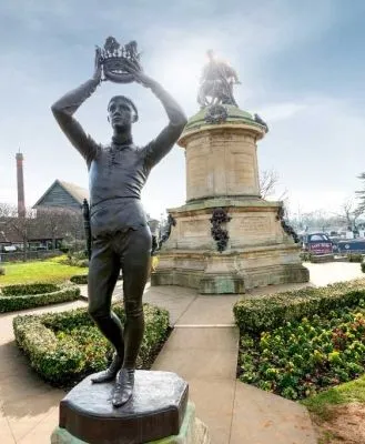 Statue of a character holding a crown in Bancroft Gardens, with the Shakespeare Memorial in the background on a sunny day.