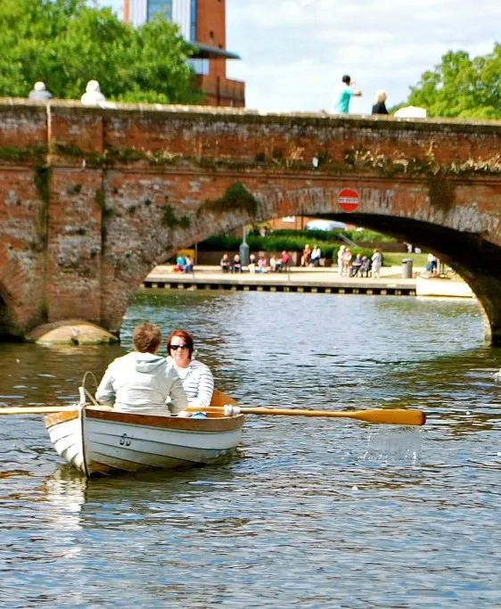 People enjoy rowing on the River Avon near a historic brick bridge, with bystanders watching from the bridge on a sunny day in Stratford-upon-Avon.