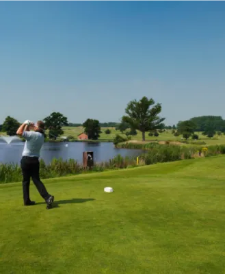 Golfer swinging on a scenic golf course with a pond and fountain in the background, surrounded by lush greenery on a bright, clear day.