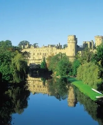 Warwick Castle's grand stone structure reflected in a calm river, surrounded by lush green trees under a clear blue sky.