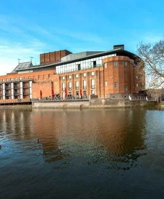 The Royal Shakespeare Company Theatre on the banks of the River Avon, a modern red-brick building with a view of the water and surrounding trees.
