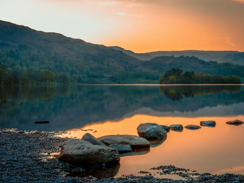 Serene lake with rocks in the foreground and hills in the background at sunset