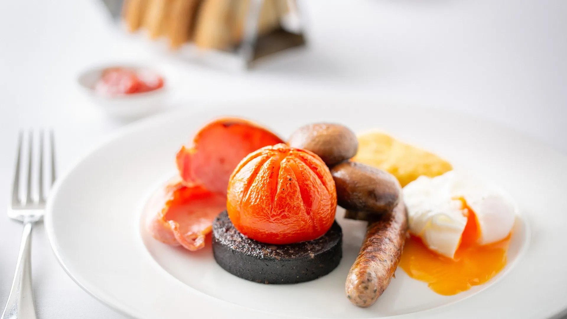Full English breakfast with sausage, black pudding, grilled tomato, mushrooms, bacon, hash brown, and poached egg on a white plate.