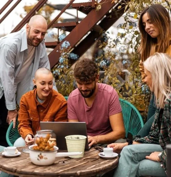 Group of six people sitting around a table outdoors, looking at a laptop and interacting with each other.