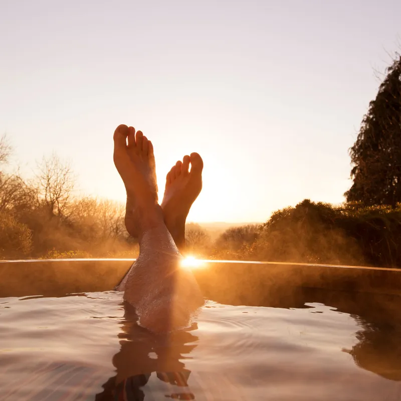 Person relaxing in an outdoor hot tub at sunset with legs crossed and steam rising from the water.