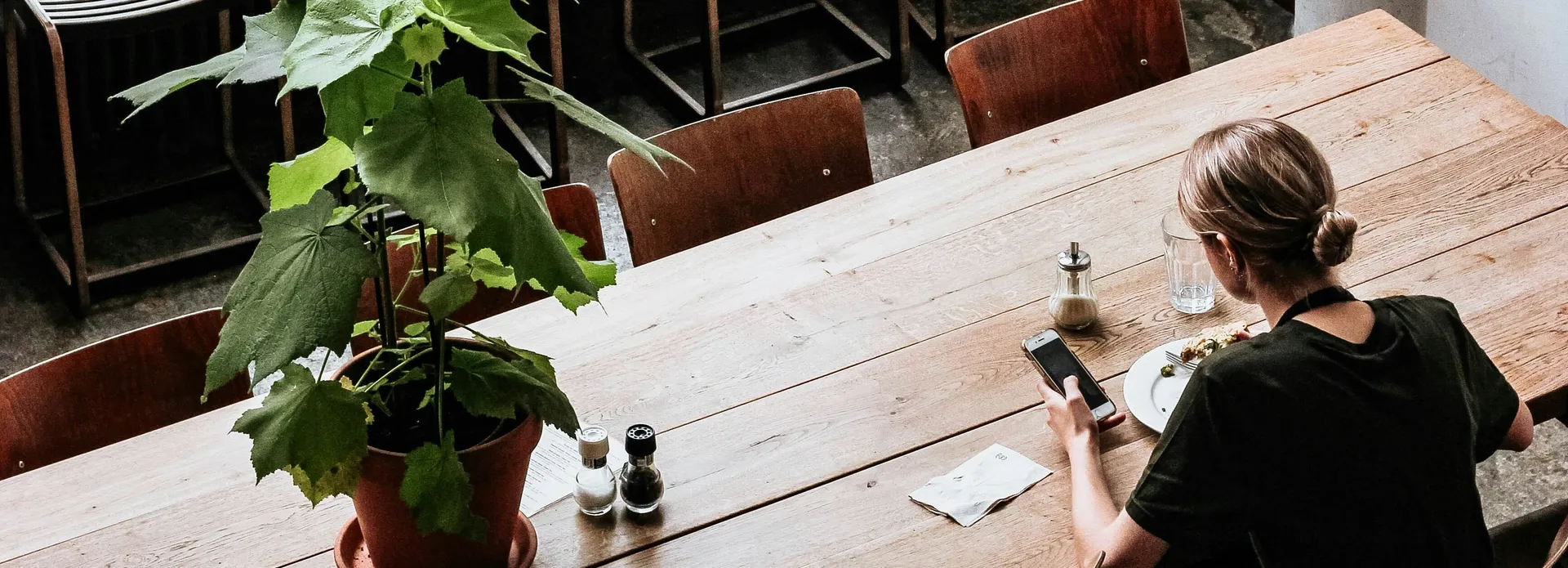 Person sitting alone at a wooden table in a modern cafe with hanging globe lights and a large plant.
