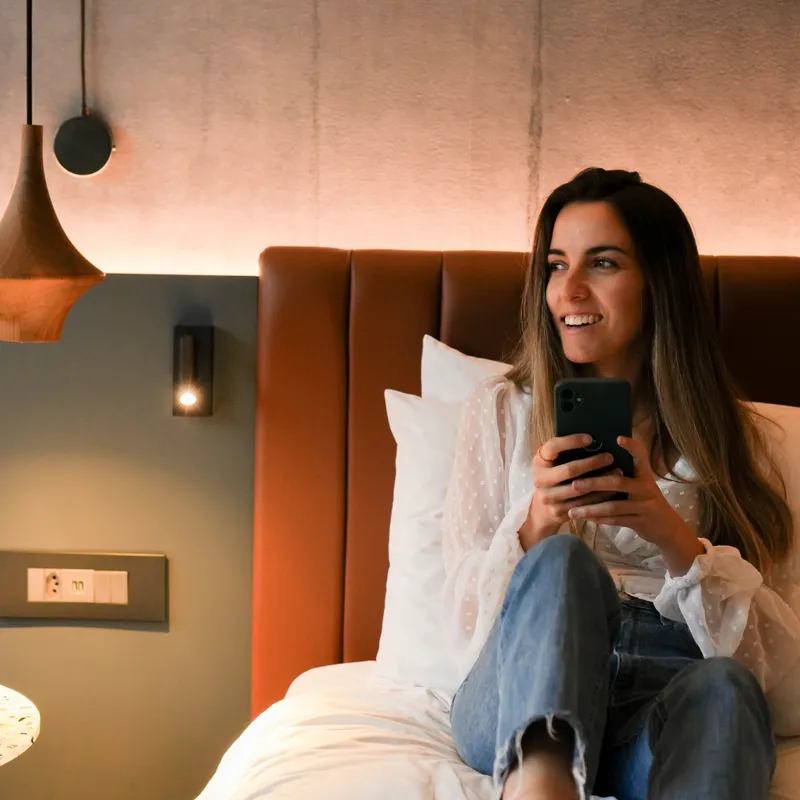 Woman sitting on bed holding a phone and smiling in a modern cozy room