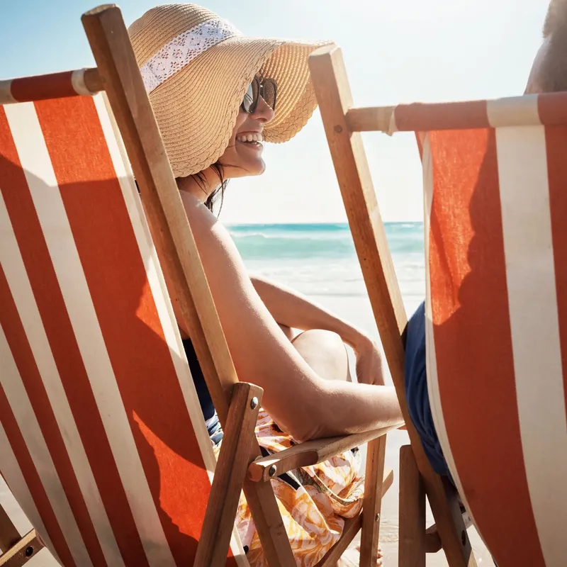 Woman wearing a sunhat and sunglasses sitting on a striped deck chair at the beach, smiling and facing a companion.