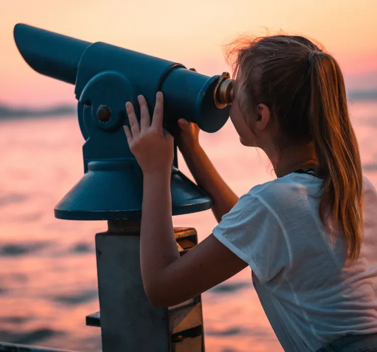 Girl looking through a blue coin-operated telescope by the water at sunset