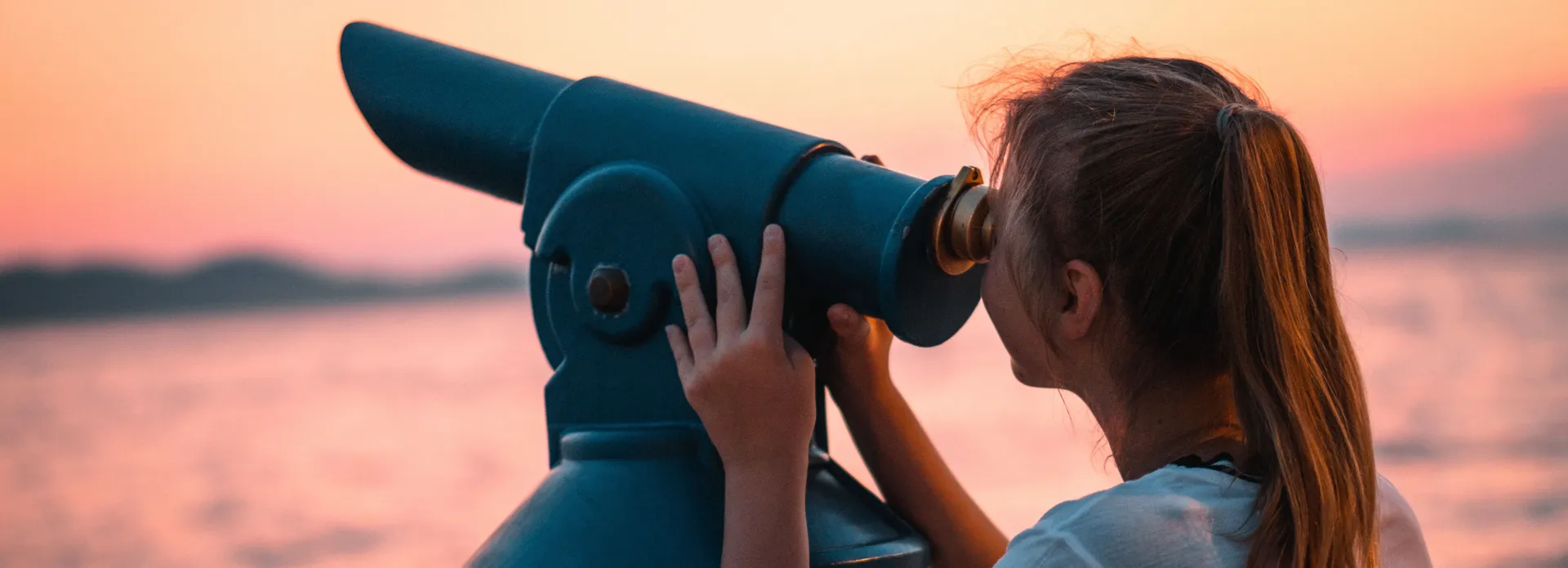 Girl looking through a blue coin-operated telescope by the water at sunset