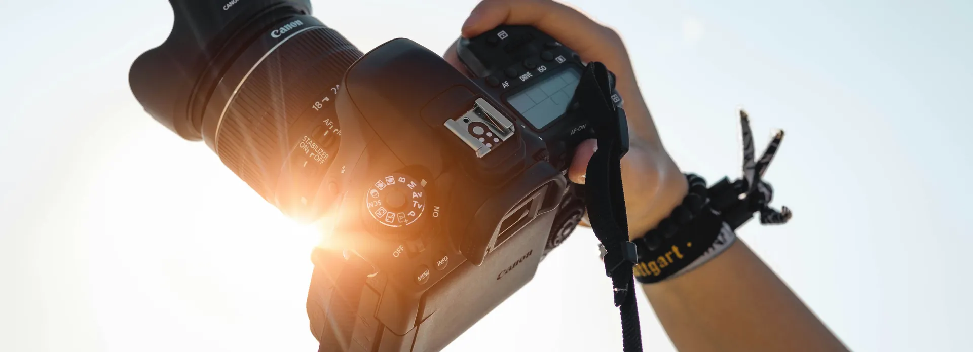 Hand holding a Canon DSLR camera against a bright sky with sunlight shining through.
