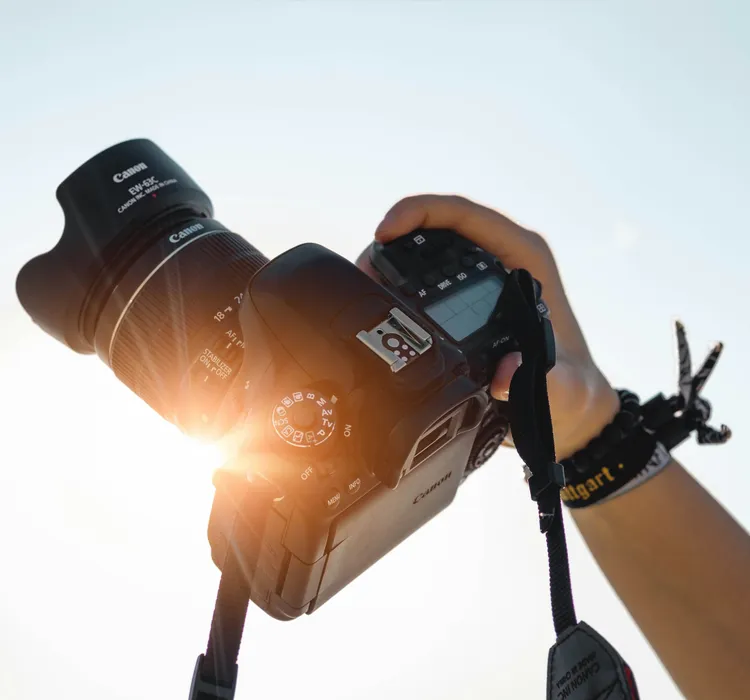 Hand holding a Canon DSLR camera against a bright sky with sunlight shining through.