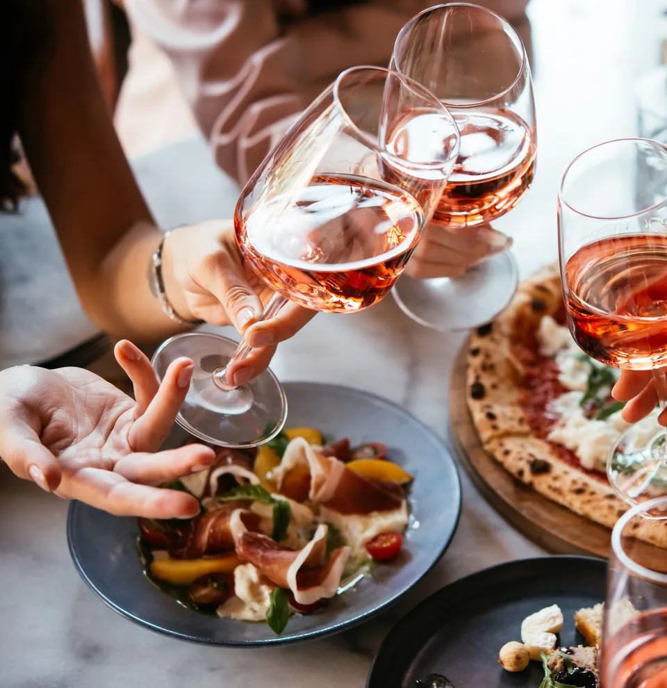 People clinking glasses of rosé wine over a table with pizza and appetizers