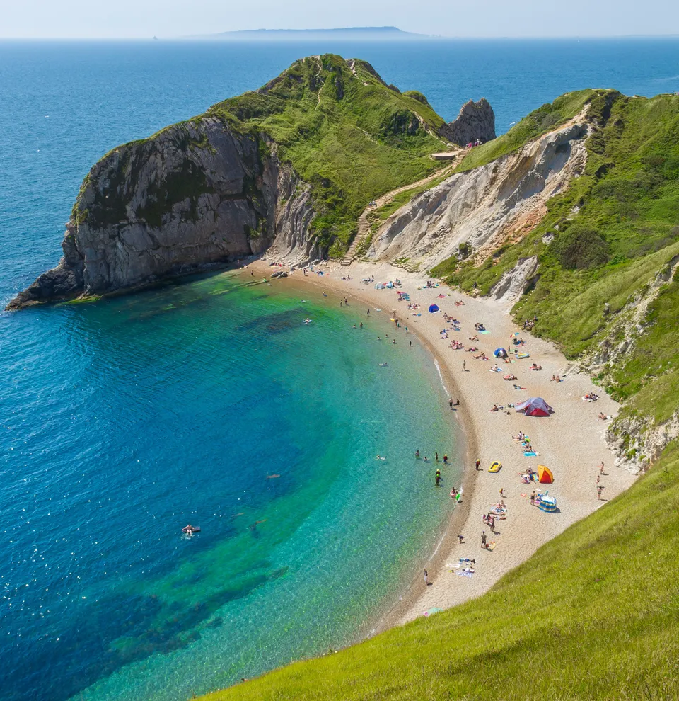 Aerial view of a curved beach with clear blue water surrounded by steep green cliffs and people sunbathing and swimming
