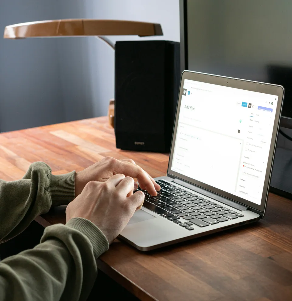 Person typing on a laptop at a wooden desk with a monitor and speaker