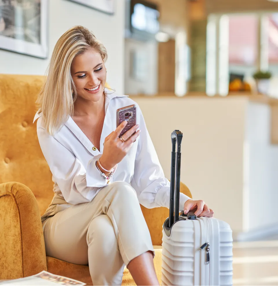 Woman sitting in a hotel lobby with a suitcase, using her smartphone