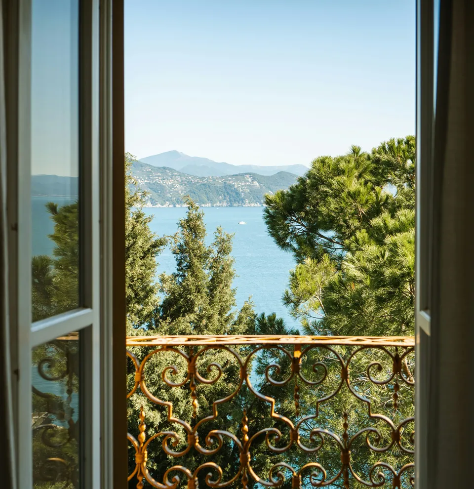 Scenic view of a blue sea and lush green trees through an open window with a decorative balcony railing.