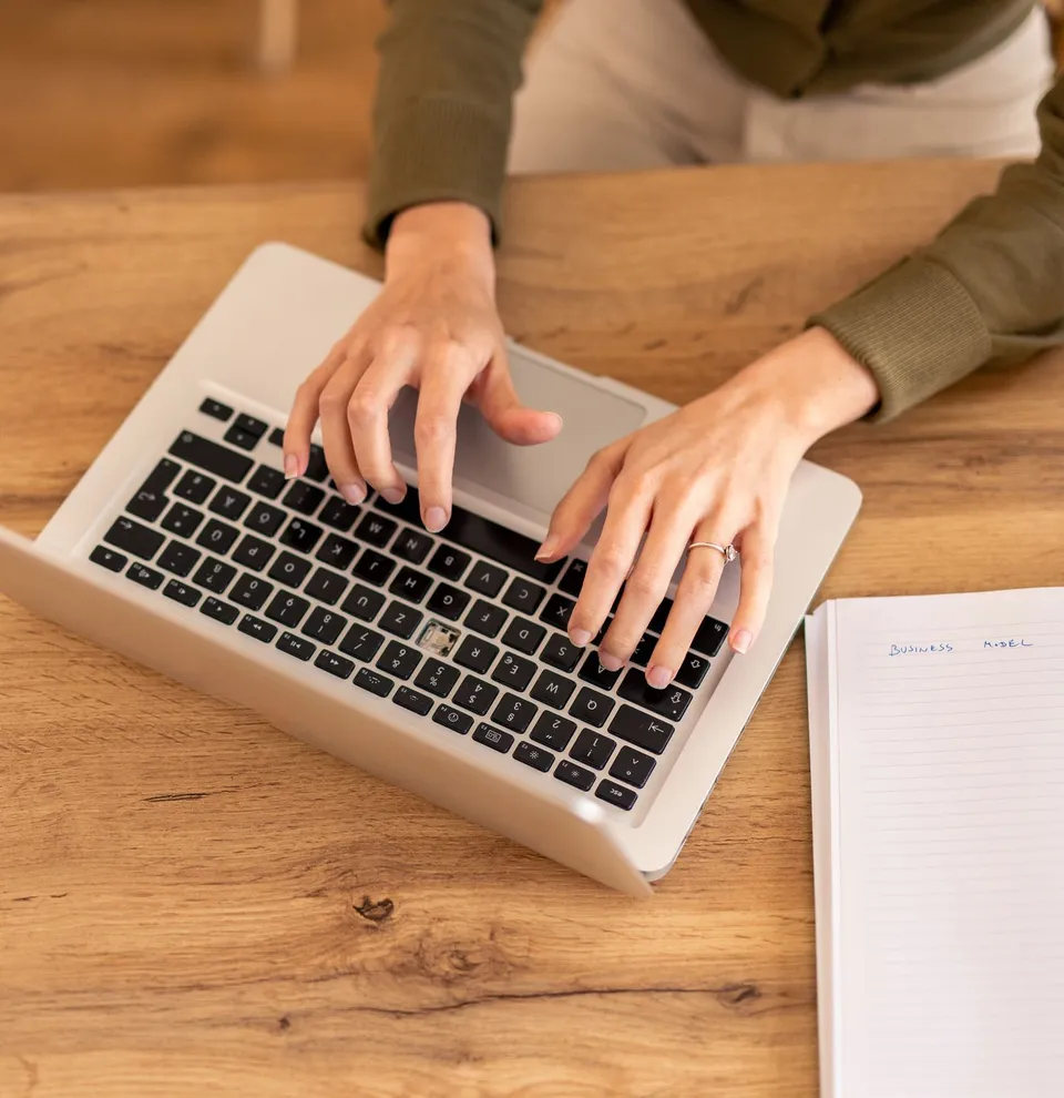 Person typing on a laptop with a notebook, smartphone, and cup on a wooden desk.