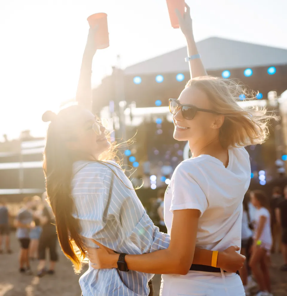 Two young women enjoying a music festival outdoors, holding drinks and having fun.