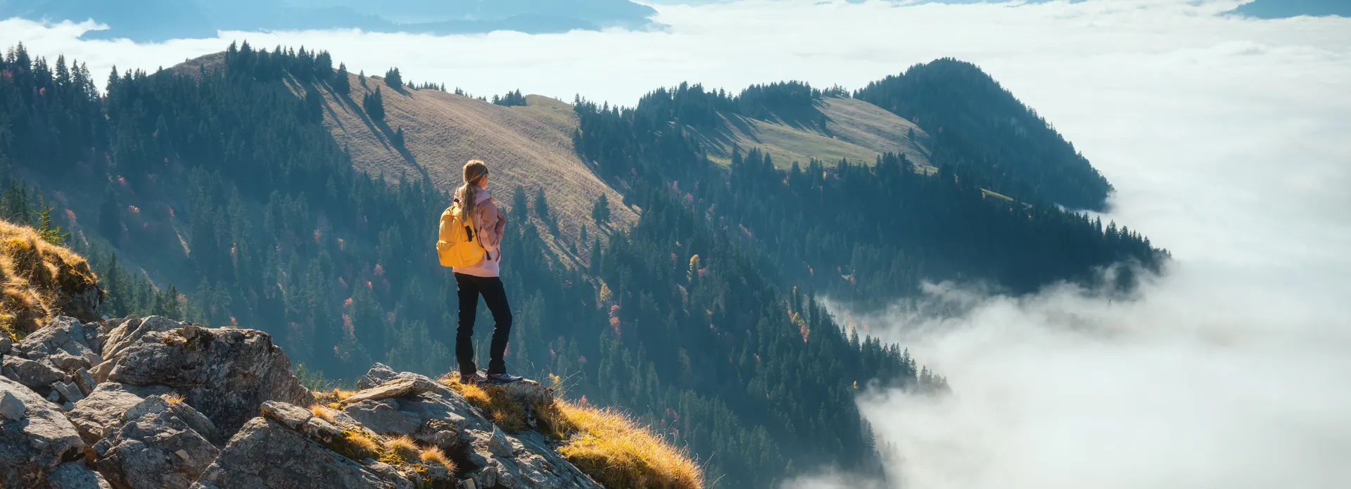 Hiker with yellow backpack standing on a rocky mountain peak overlooking a misty forest landscape