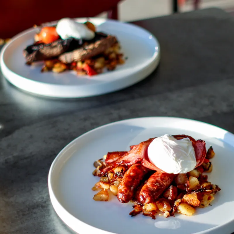 Two plated cooked breakfasts with sausages, bacon, fried potatoes and poached eggs beside glasses of orange juice.