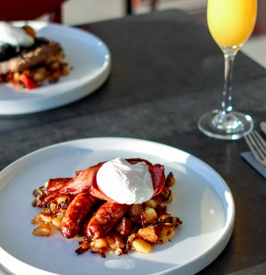 Two plated cooked breakfasts with sausages, bacon, fried potatoes and poached eggs beside glasses of orange juice.