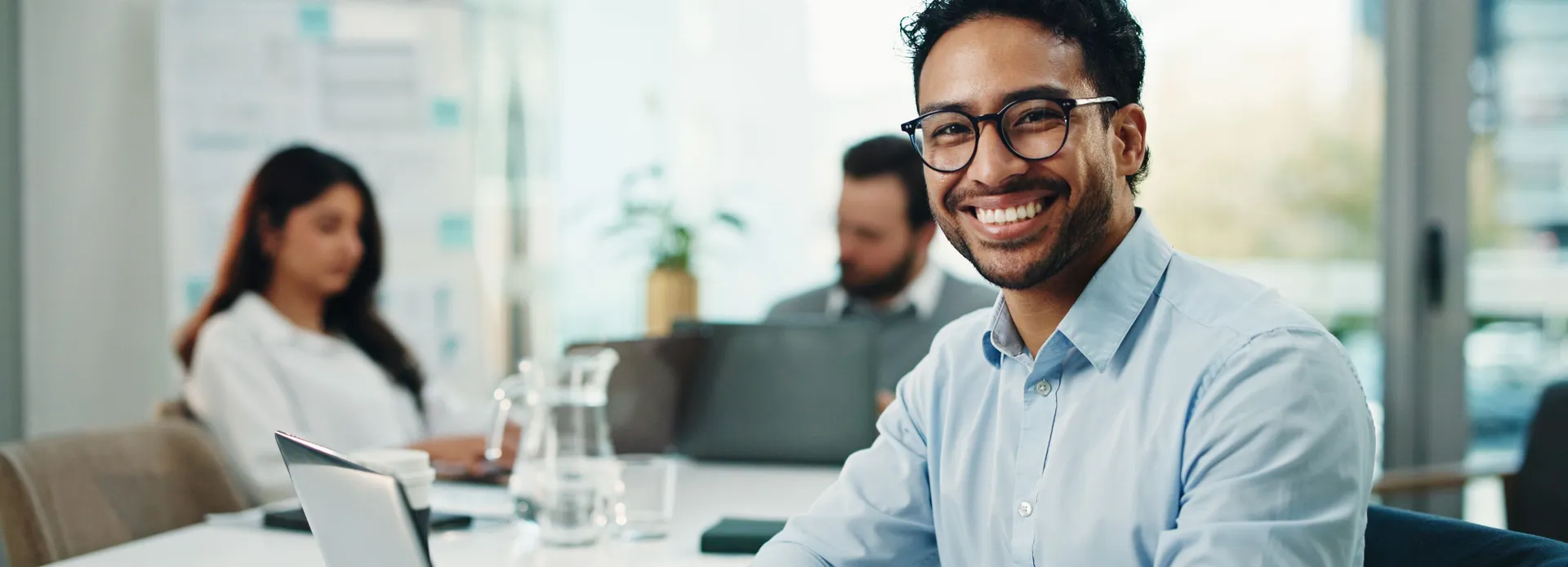 Smiling man wearing glasses seated at a laptop in a bright office, with two colleagues working behind him