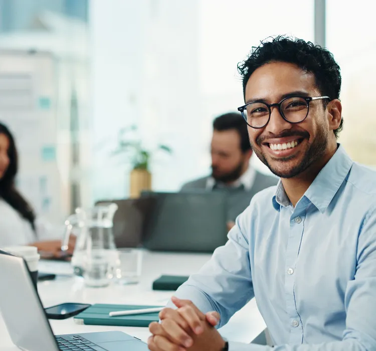 Smiling man wearing glasses seated at a laptop in a bright office, with two colleagues working behind him
