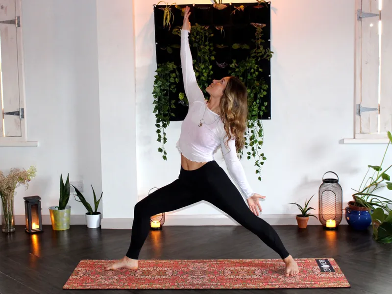 Person practicing yoga indoors in a warrior pose with plants and candles in the background.