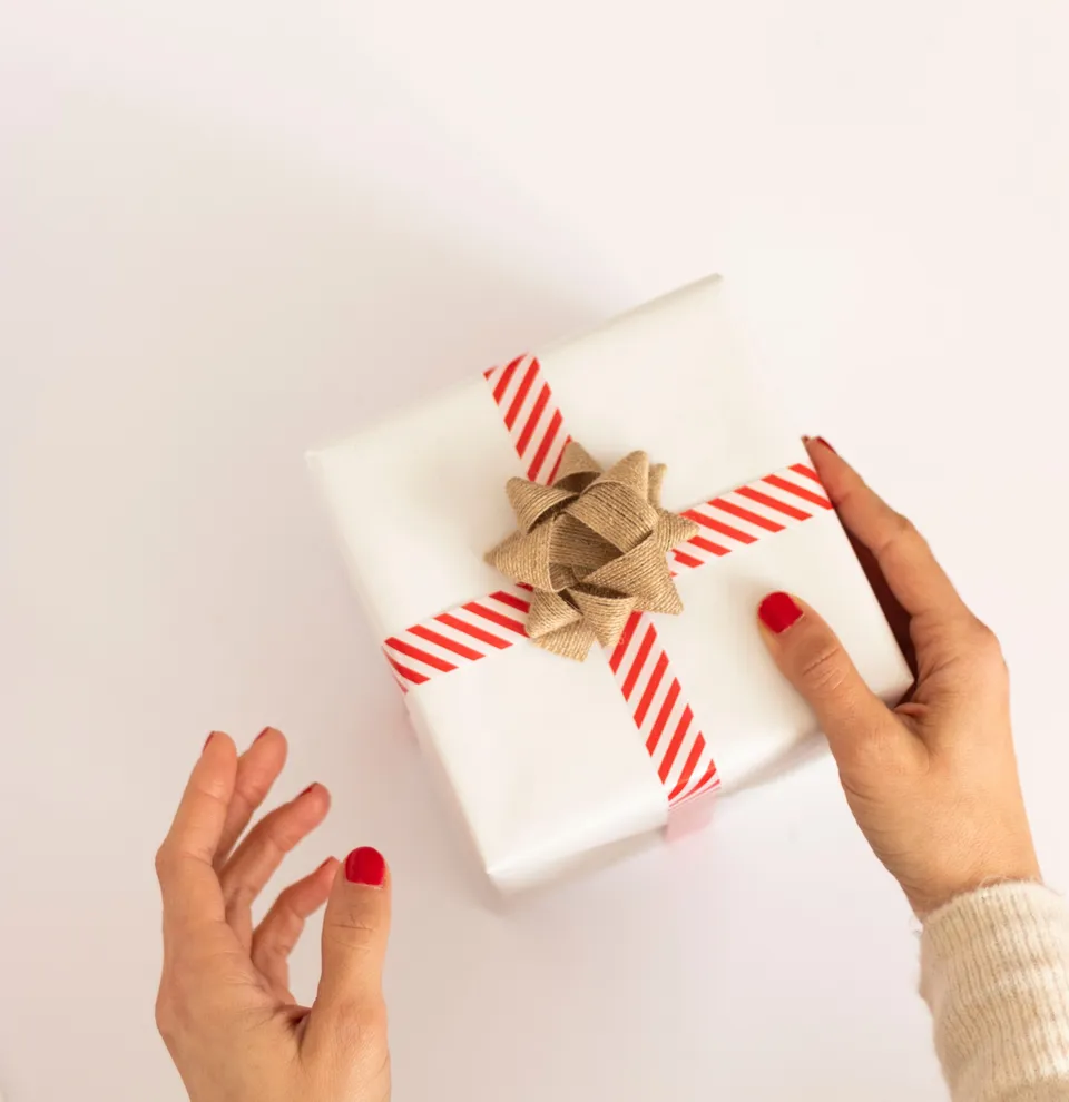 Hands holding a white gift box with red striped ribbon and a burlap bow