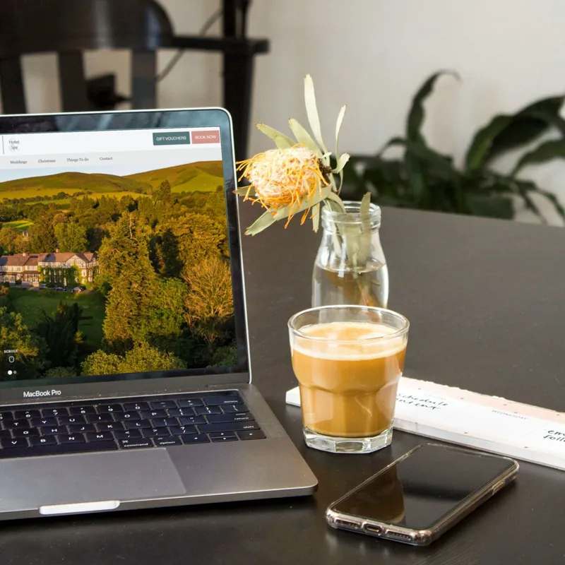 Workspace with laptop, coffee, flower in vase, and phone on a table.