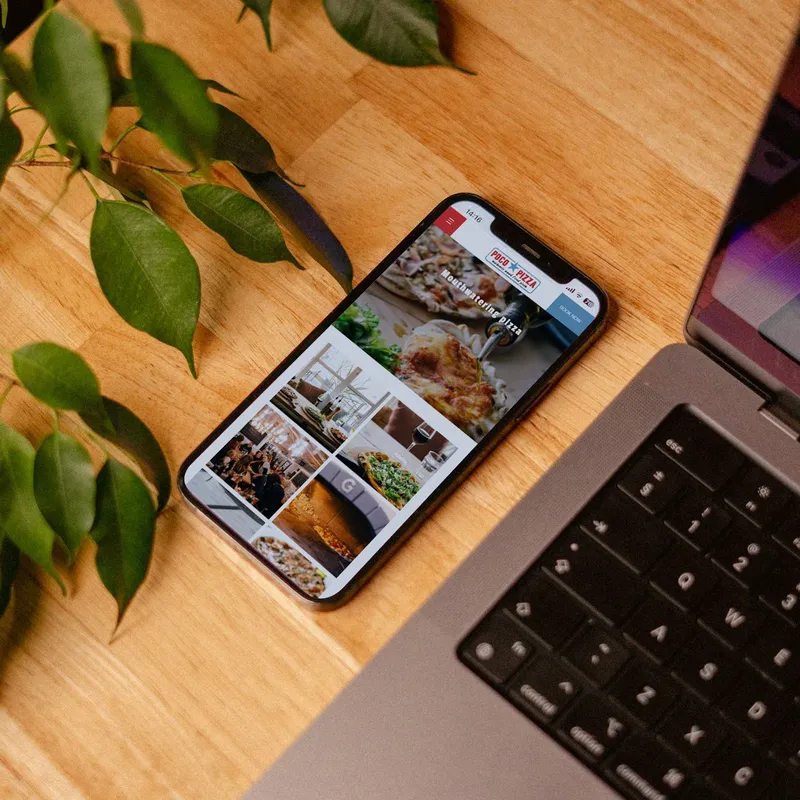 Smartphone displaying a pizza restaurant website next to a laptop on a wooden desk