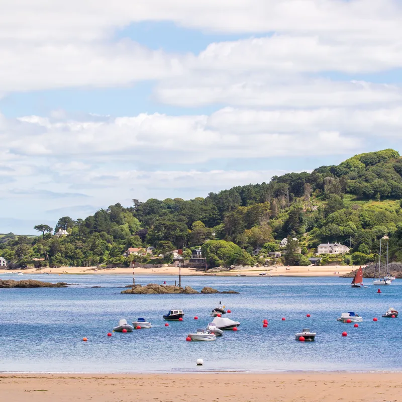 Small boats floating on calm water near a sandy beach with green hills and houses in the background