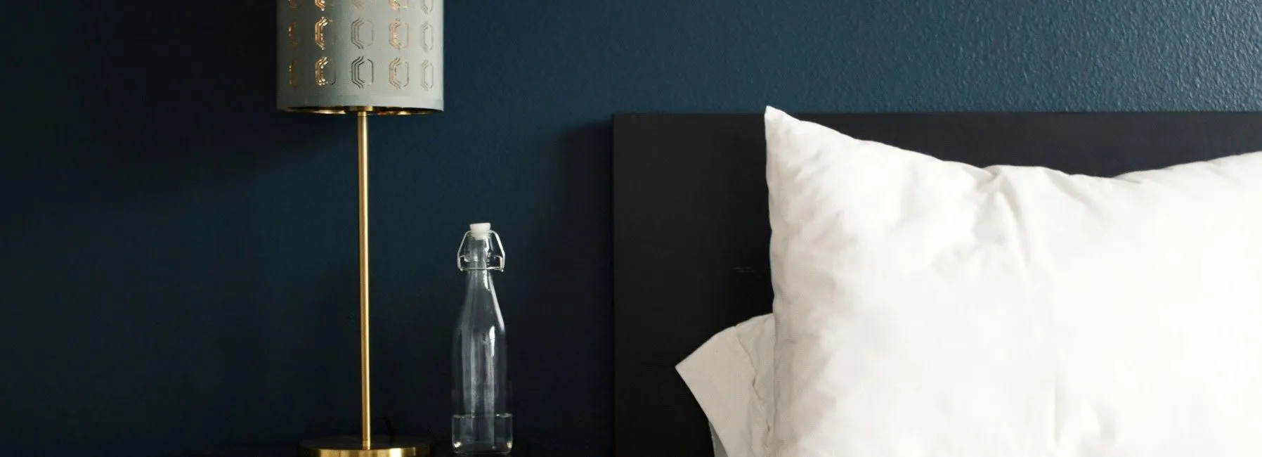 Modern bedroom setting with a gold lamp, glass water bottle, white pillow, and dark blue wall.
