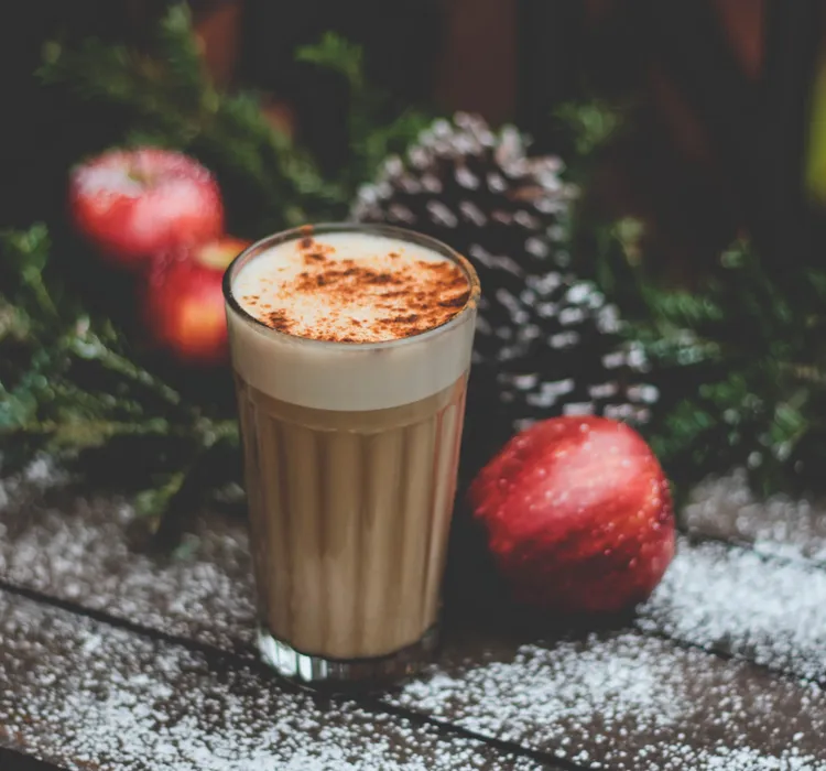 A glass of frothy coffee topped with cinnamon surrounded by apples, pine cones, and evergreen branches on a wooden surface dusted with powdered sugar.