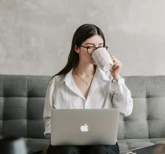 Woman sitting on a couch using a laptop and drinking from a mug
