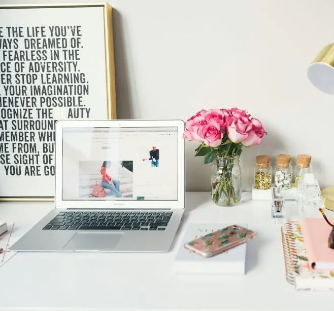 Organized workspace with a laptop, pink flowers in a jar, gold desk lamp, and notebooks on a white desk
