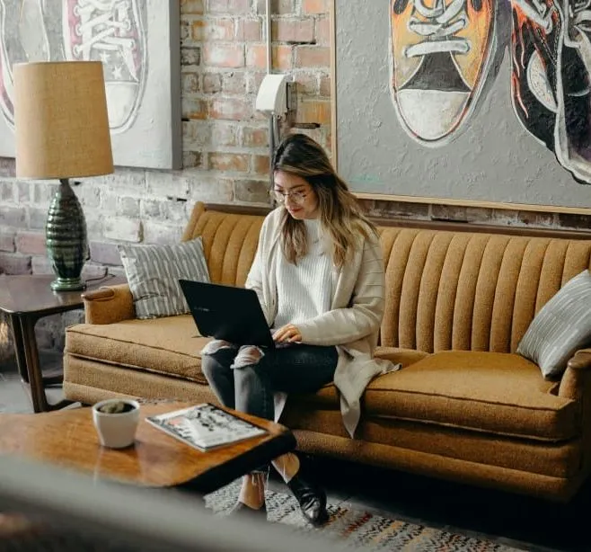 Woman sitting on a brown sofa working on a laptop in a stylish living room with brick walls and art on the wall.