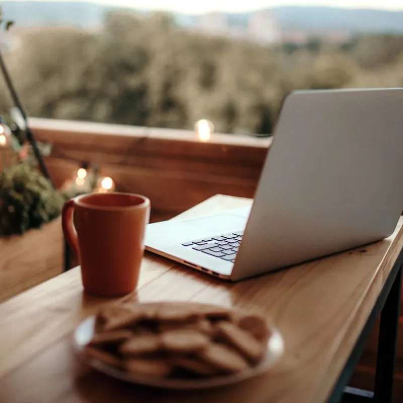 Laptop, coffee mug, and plate of cookies on a wooden table on a cozy balcony with string lights.