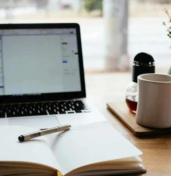 Laptop, open notebook with pen, coffee mug, and flowers on a desk
