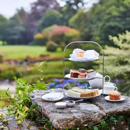 Afternoon tea setup on a stone surface outdoors, with sandwiches, scones, desserts, and a teapot against a lush garden backdrop.
